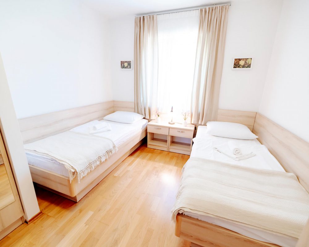 Das Zweibettzimmer verfügt über ein großes Fenster zwischen den zwei komfortablen Einzelbetten. Vor den Betten sind zwei kleine Teppiche.