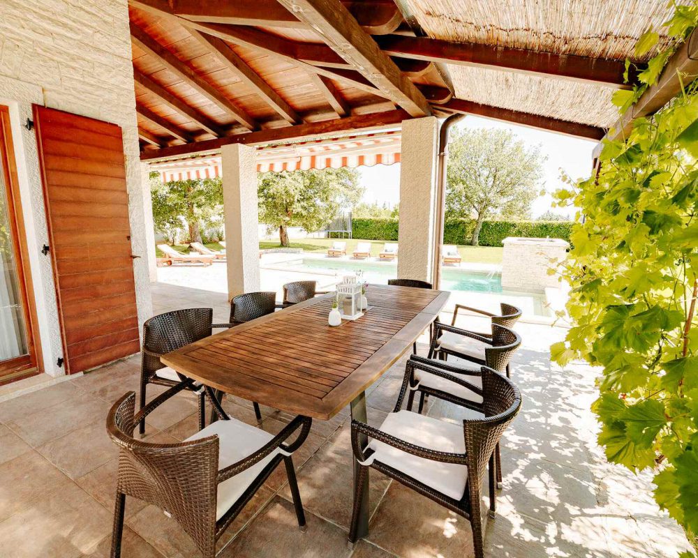 Auf der überdachten Terrasse steht ein langer hübscher Esstisch mit acht Sitzmöglichkeiten. Am Rand der Terrasse wachsen Weinstöcke.